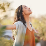 Young latin woman with arms outstretched breathing in fresh air during sunrise at the balcony. Healthy girl enjoying nature while meditating during morning with open arms and closed eyes. Mindful woman enjoying morning ritual while relaxing in outdoor park.