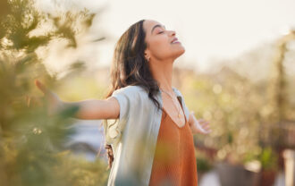 Young latin woman with arms outstretched breathing in fresh air during sunrise at the balcony. Healthy girl enjoying nature while meditating during morning with open arms and closed eyes. Mindful woman enjoying morning ritual while relaxing in outdoor park.