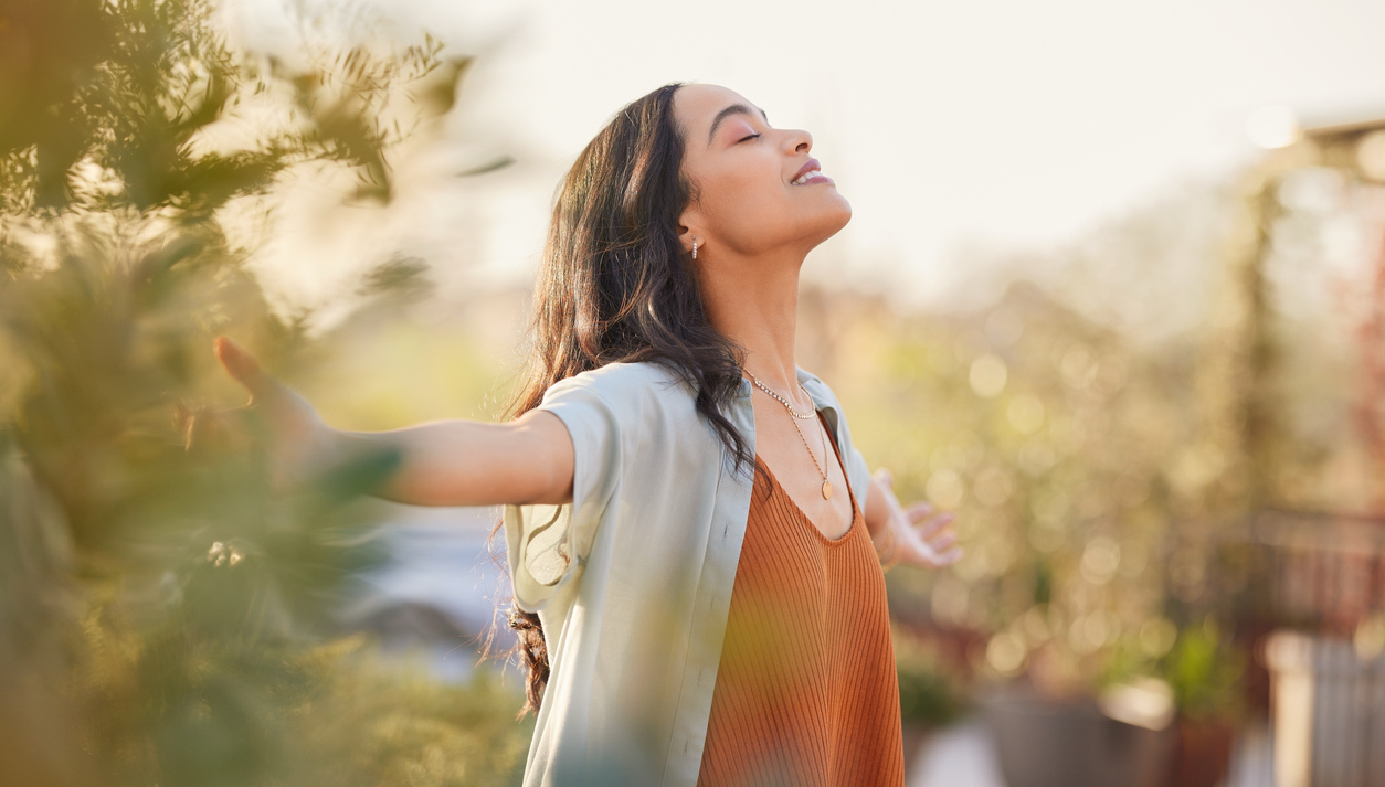 Young latin woman with arms outstretched breathing in fresh air during sunrise at the balcony. Healthy girl enjoying nature while meditating during morning with open arms and closed eyes. Mindful woman enjoying morning ritual while relaxing in outdoor park.